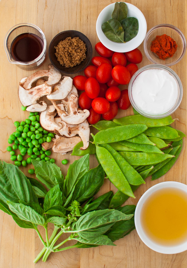 Snow peas, tomatoes, Thai basil, mushrooms, peas, kaffir lime leaves, curry paste, and coconut milk on a wooden cutting board.