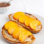 Peach slices on toasted bread on a white plate next to a bowl of honey.