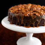 A chocolate cake topped with caramel sauce and toasted pecans on a white cake stand.