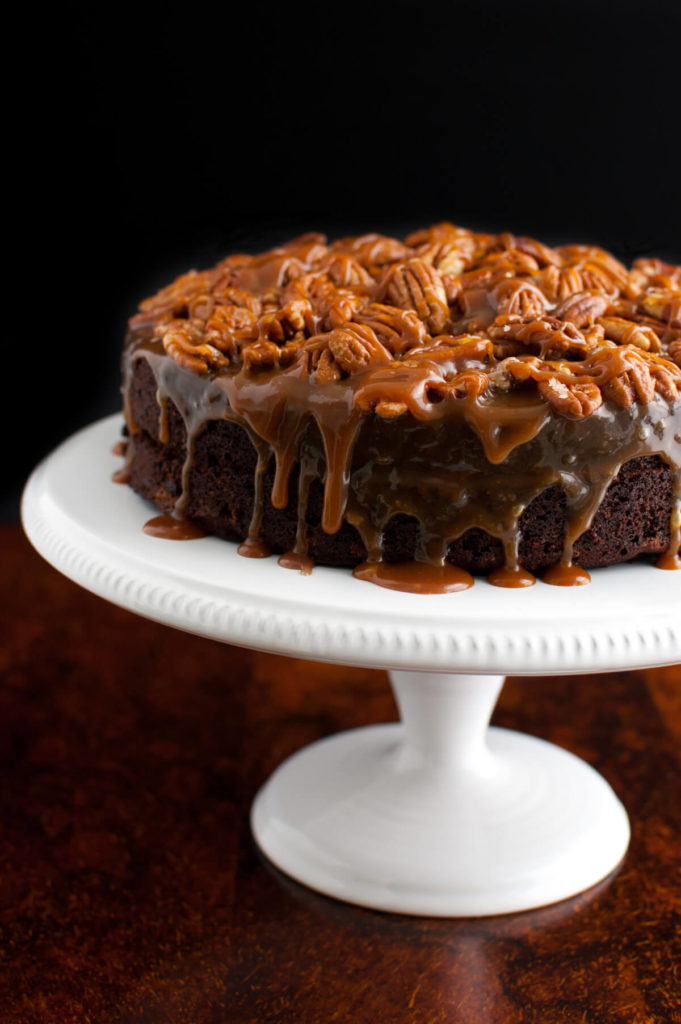 A chocolate cake topped with caramel sauce and toasted pecans on a white cake stand.
