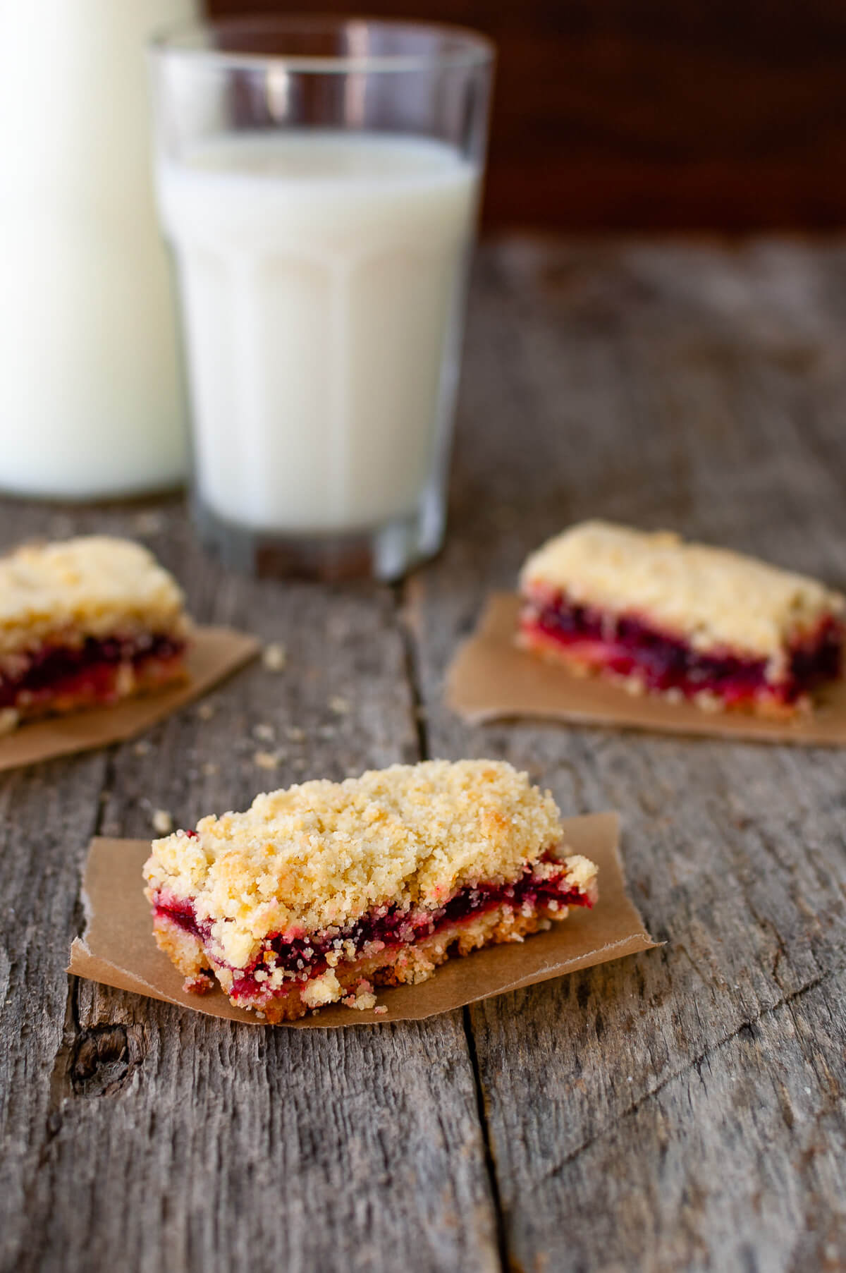 Some cranberry orange crumb bars on pieces of parchment paper next to a glass of milk.