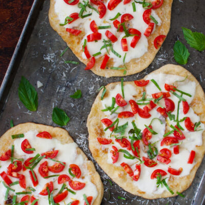 Three small pizzas topped with cheese and fresh basil and tomatoes on a metal tray.