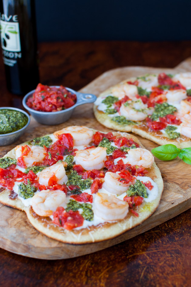 Shrimp pizzas on a wooden cutting board next to bowls of pesto and tomato sauce.