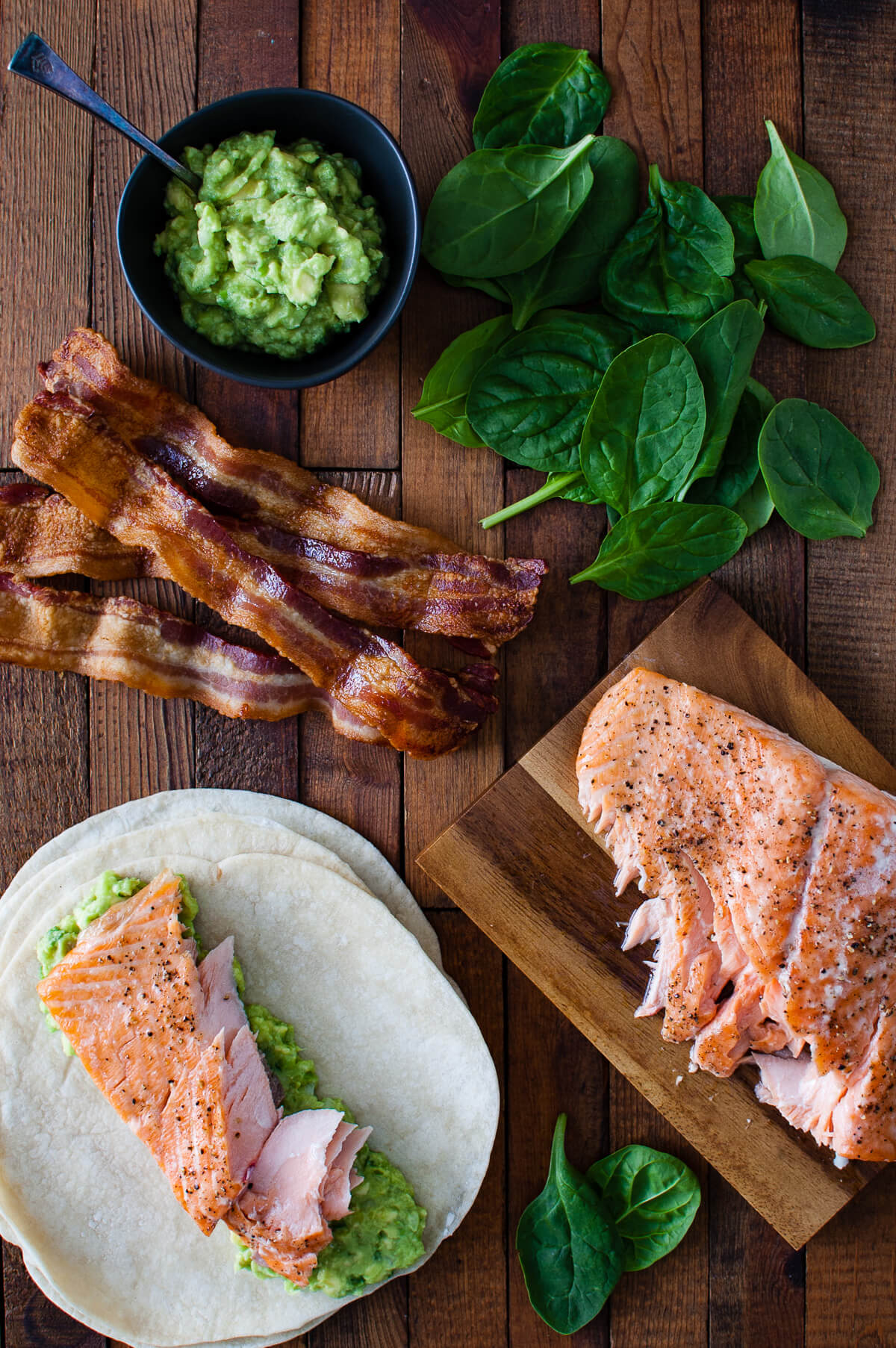 Roasted salmon, guacamole in a bowl, spinach leaves, crispy bacon, and tortillas laid out on a wooden cutting board.