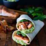 Salmon, guacamole, bacon, and spinach wrapped inside a tortilla on a wooden board.