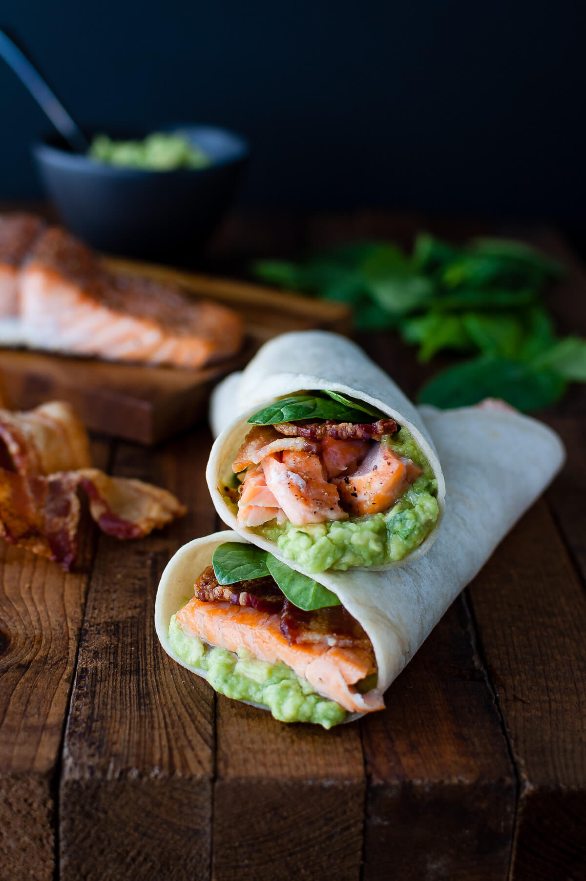 Salmon, guacamole, bacon, and spinach wrapped inside a tortilla on a wooden board.