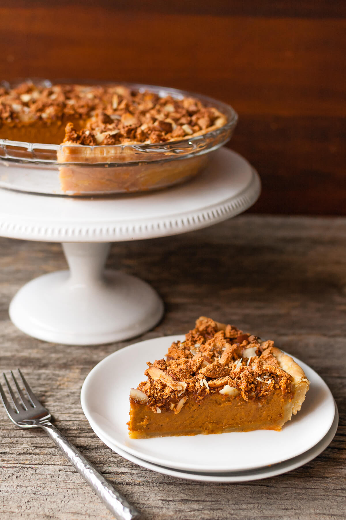 A slice of pumpkin pie on a plate next to a pie on a white cake stand.