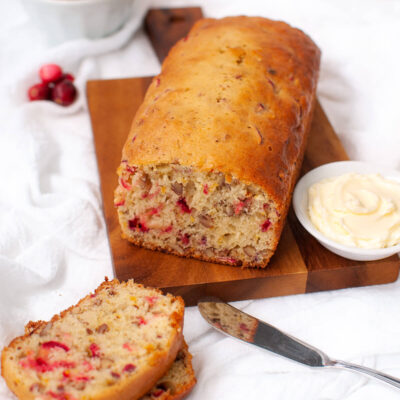 Loaf of cranberry bread on wooden board next to slices of bread and dish of softened butter.