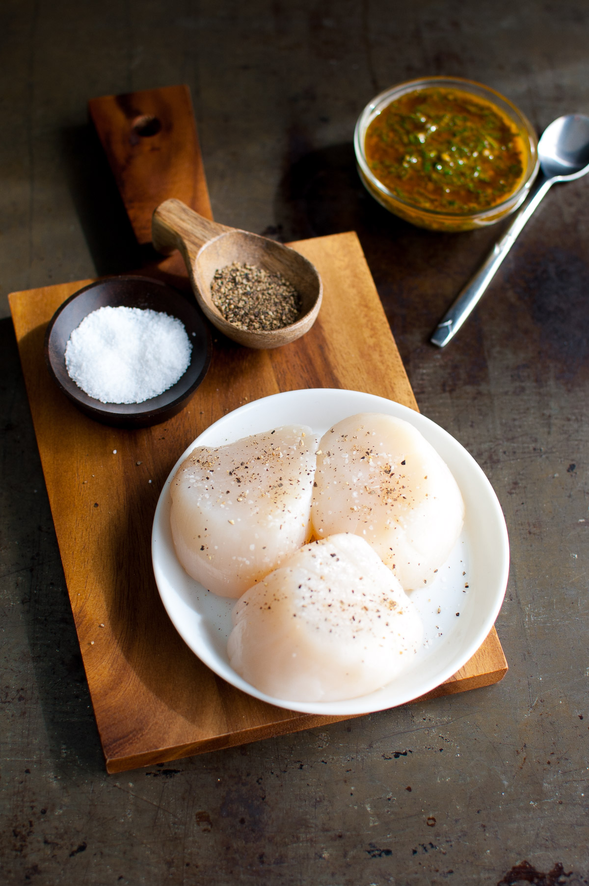 Raw scallops on a plate next to pinch bowls of salt and pepper.