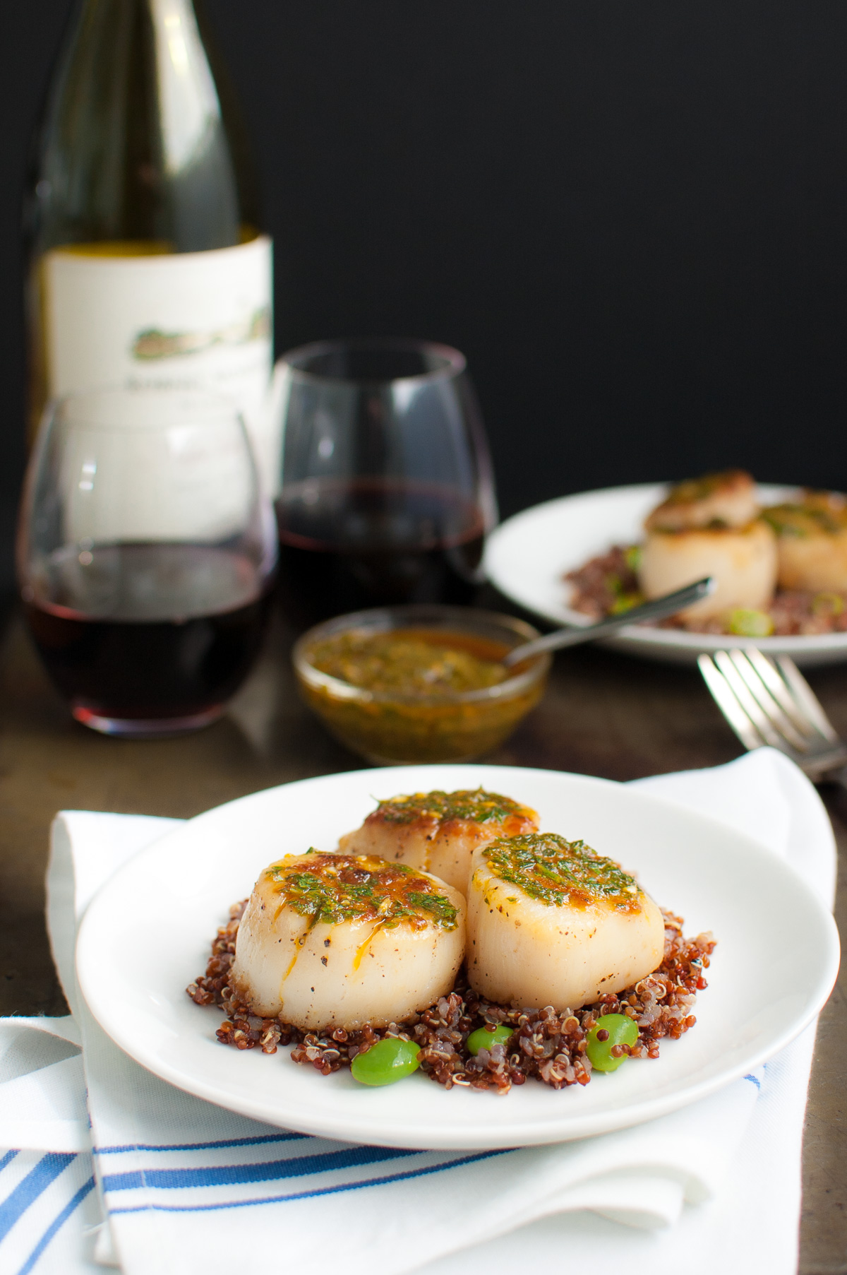 Two plates of seared scallops, a bowl of chermoula sauce, and some glasses of wine.