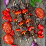 Skewers of beef kebabs next to some grilled tomatoes and onions.