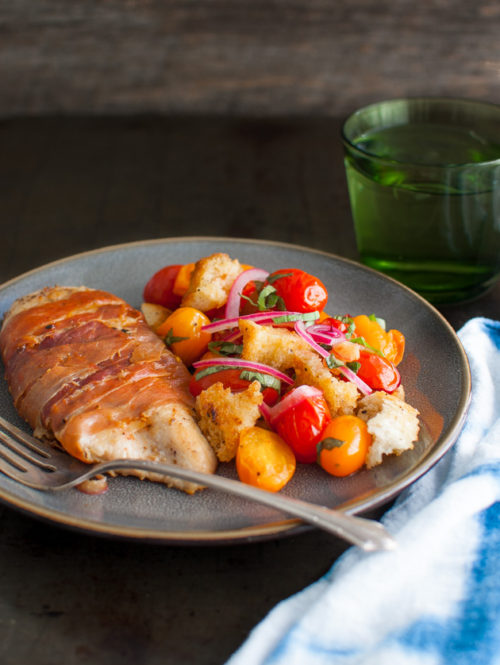 Prosciutto wrapped chicken cutlets next to a tomato, bread, and onion salad.