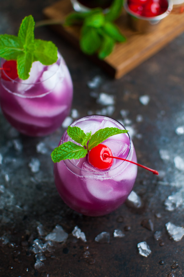 Two glasses of hibiscus tea cocktail garnished with mint and a maraschino cherry.