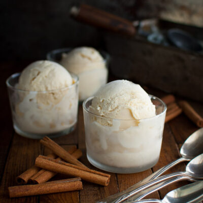 Scoops of cinnamon ice-cream in glass dessert cups next to some cinnamon sticks.