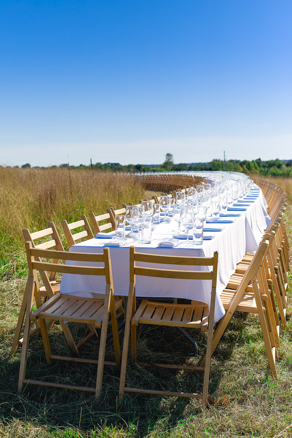 A long curved table set with white napkins and wine glasses in a field.