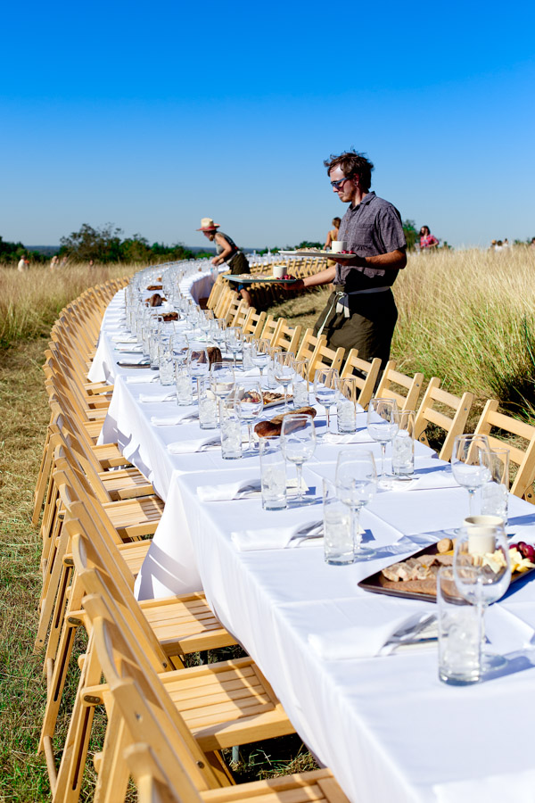 Servers setting platters of food on a long table in a field.