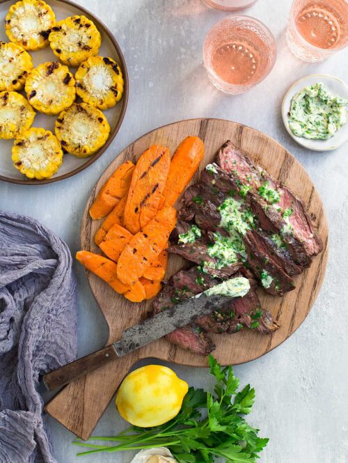 Steak slices topped with parsley-lemon-garlic butter on a cutting board and served with grilled vegetables.