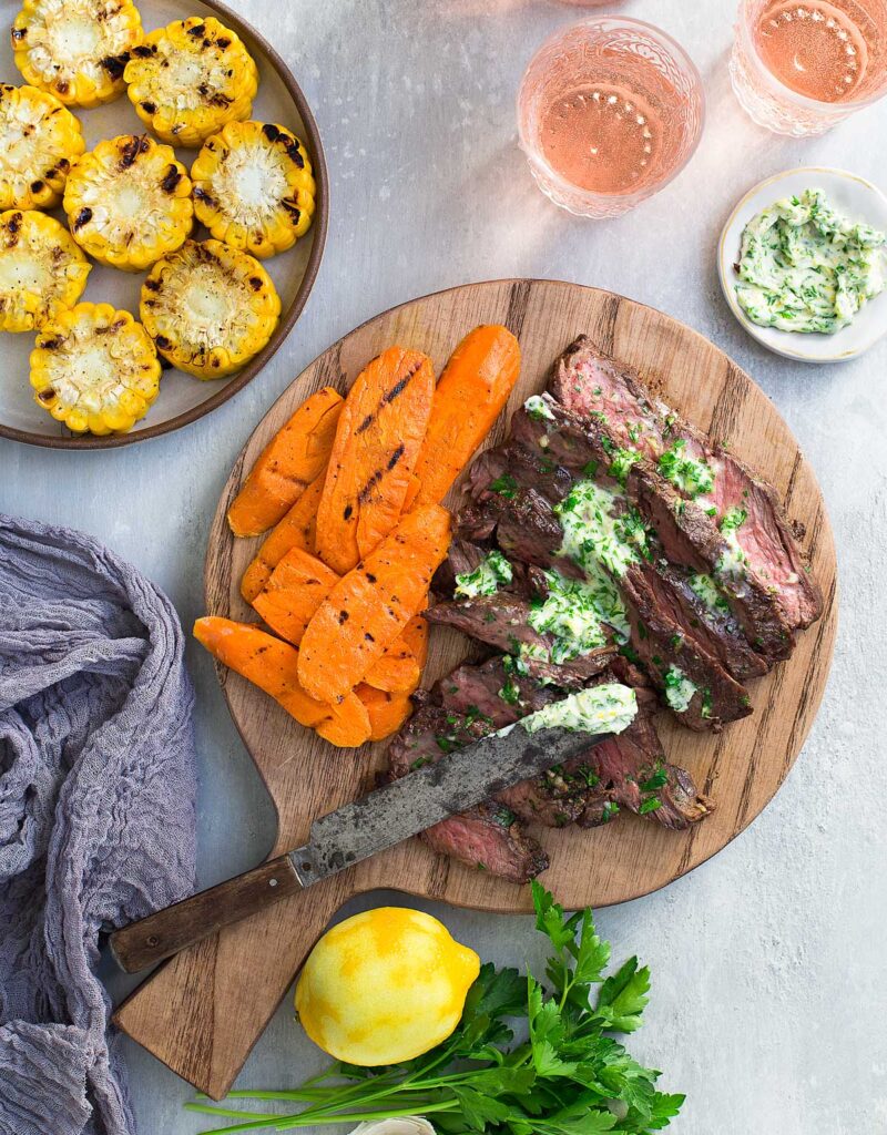 Steak slices topped with parsley-lemon-garlic butter on a cutting board and served with grilled vegetables.