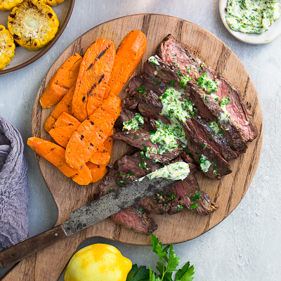 Steak slices topped with parsley-lemon-garlic butter on a cutting board and served with grilled vegetables.