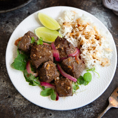 Shaking beef on a white plate served with steamed white rice, watercress, and lime wedges.