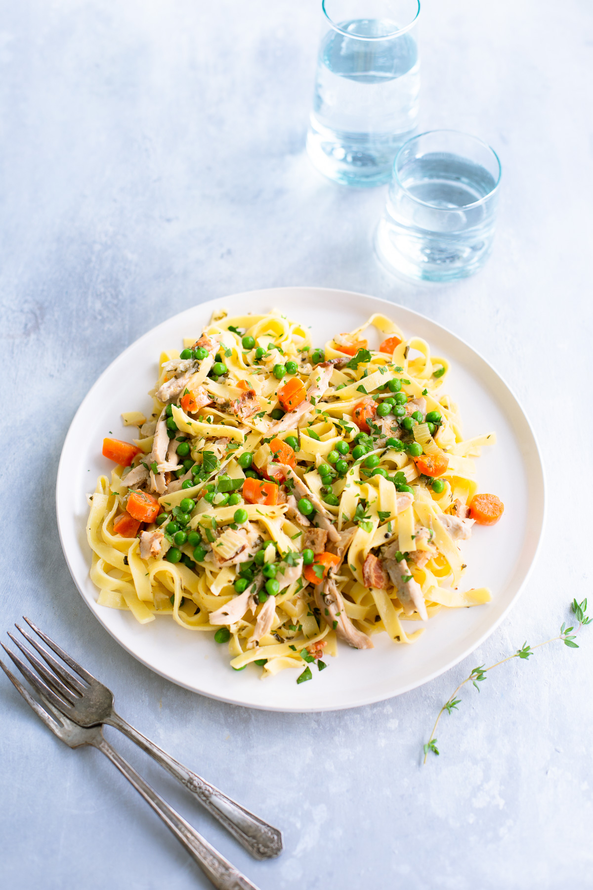 Fettuccine with chicken, peas, and carrots on a white plate next to a fork and glasses of water.