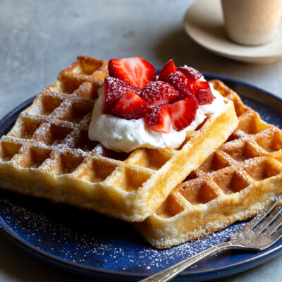 Two Belgian waffles topped with whipped cream and strawberries on a blue plate.
