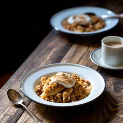 Two plates of breakfast apple crisp and a cup of coffee on a wooden table.