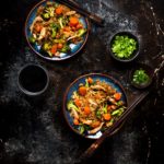 Vegetable and chicken fried rice on two blue plates with wooden chopsticks shown with a small bowl of chopped green onions, a small bowl of chopped cilantro and a cup of tea.