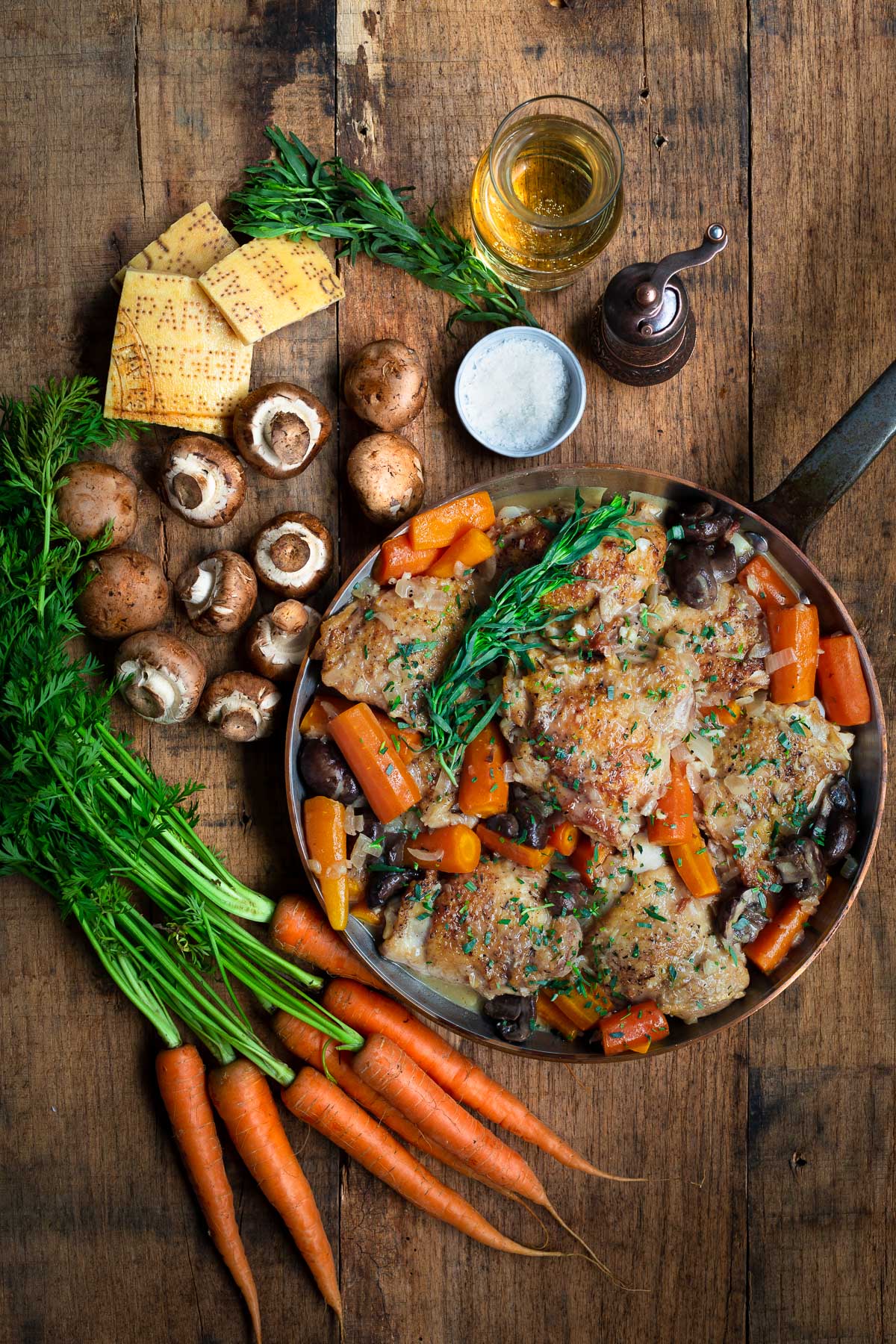 Braised chicken and carrots in a pan on a wooden surface with white wine, fresh carrots, mushrooms, tarragon, and Parmesan cheese rinds.