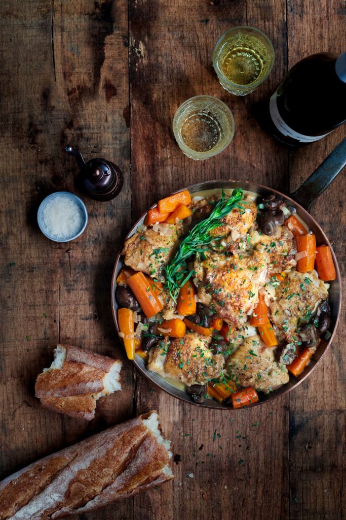 Braised chicken and carrots in a pan on a wooden surface with white wine and a baguette.
