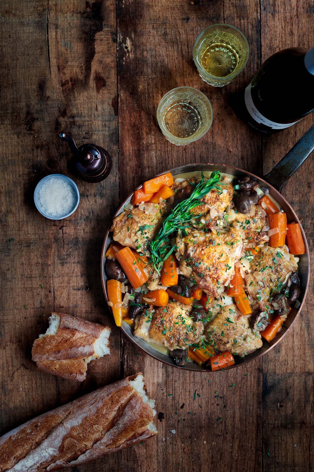 Braised chicken and carrots in a pan on a wooden surface with white wine and a baguette.