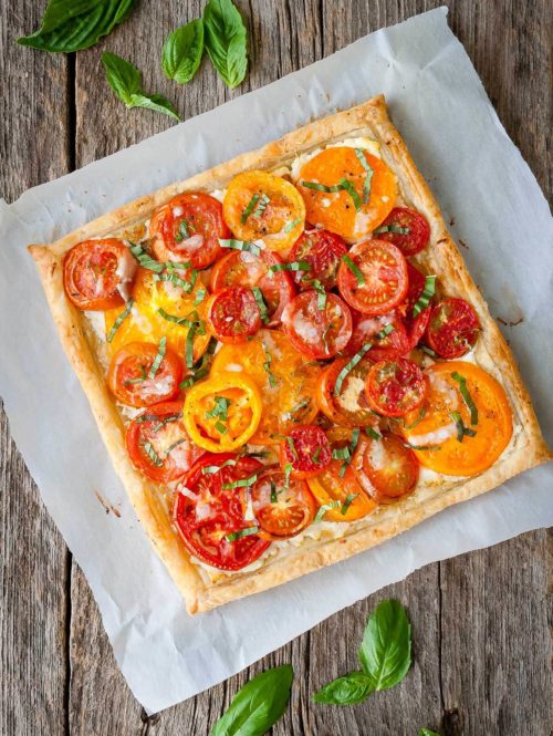 Tomato tart with goat cheese and caramelized onions on a piece of parchment paper with loose basil leaves.