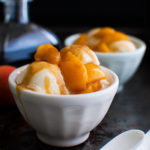 Two bowls of ice-cream topped with peaches foster shown with two white spoons and some peaches and a bottle of rum in the background.