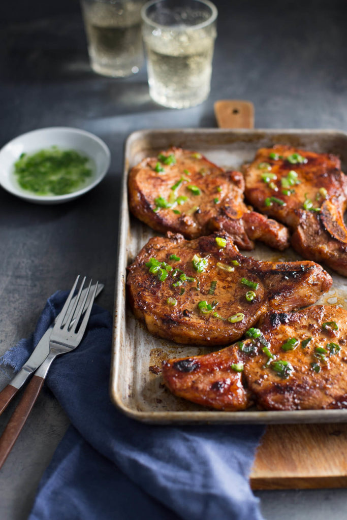 Sheet pan of pork chops with blue napkin, fork, and knife in the front and dish of scallion oil and glasses of white wine in the background.