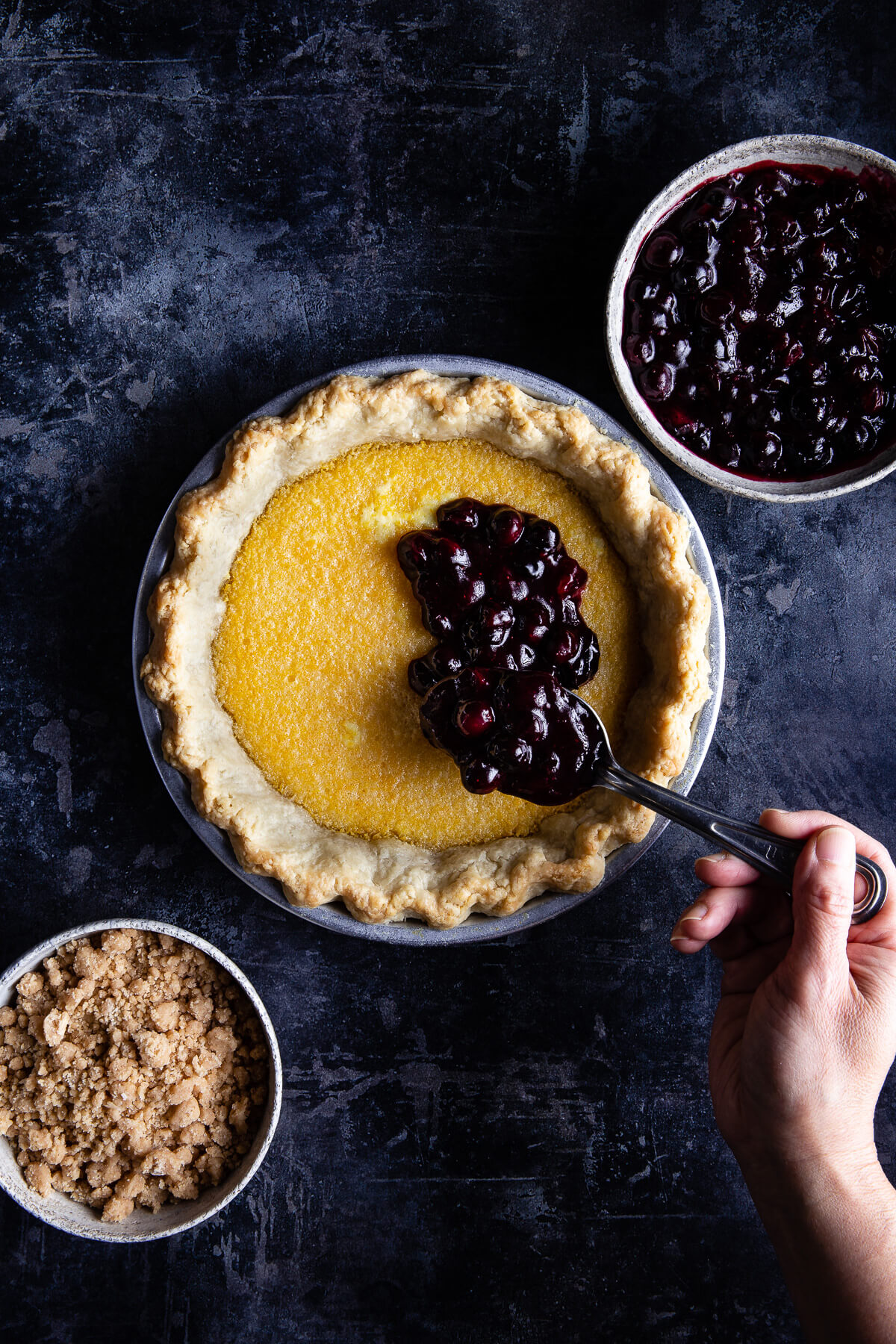 Someone spooning blueberry filling on to the lemon filling of a lemon blueberry pie.