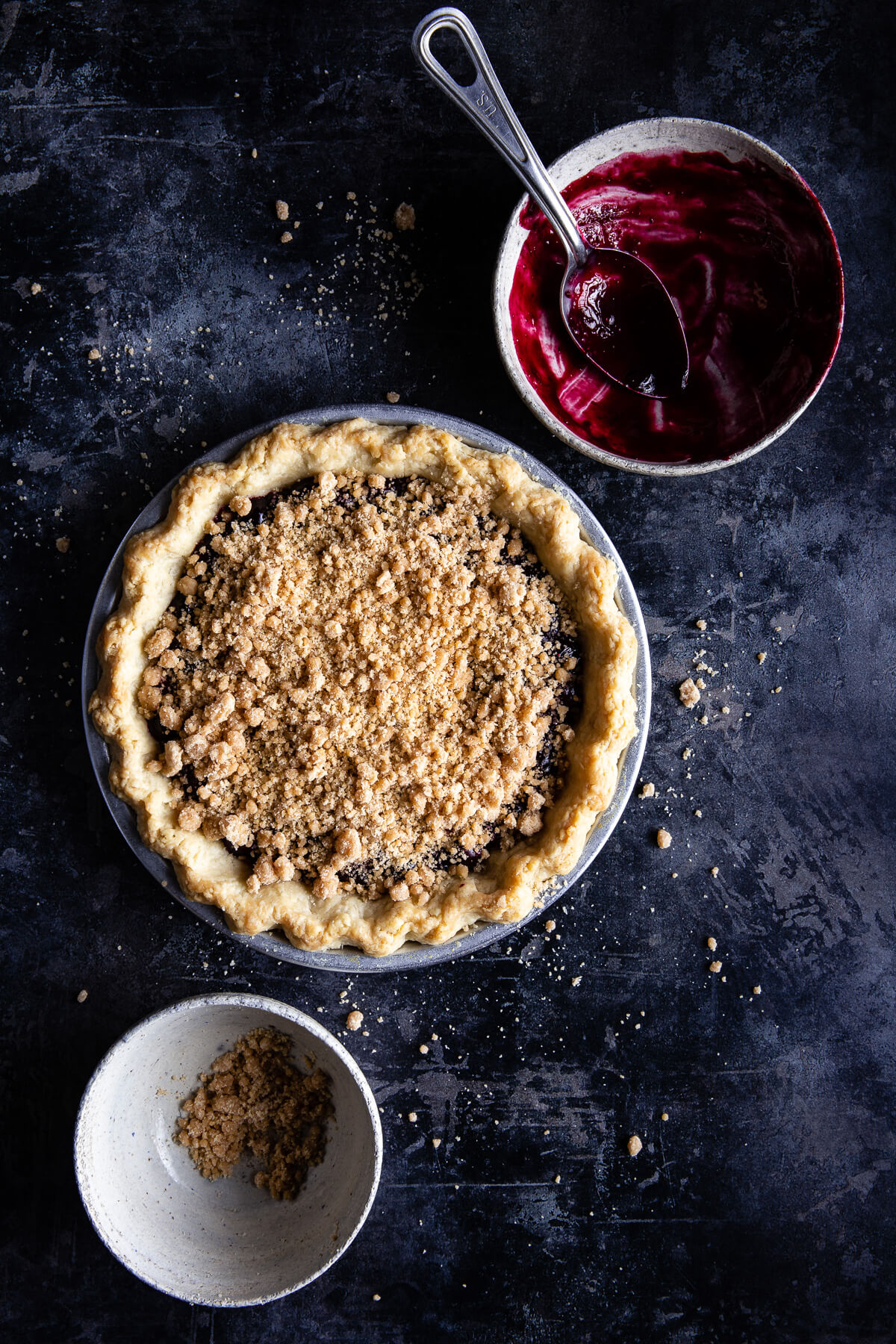 A blueberry crumb pie next to empty bowls of blueberry filling and streusel topping.