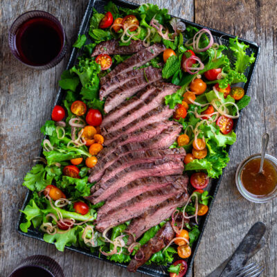 Overhead view of sliced Thai beef with salad greens, tomatoes, and, fresh herbs on a metal tray.