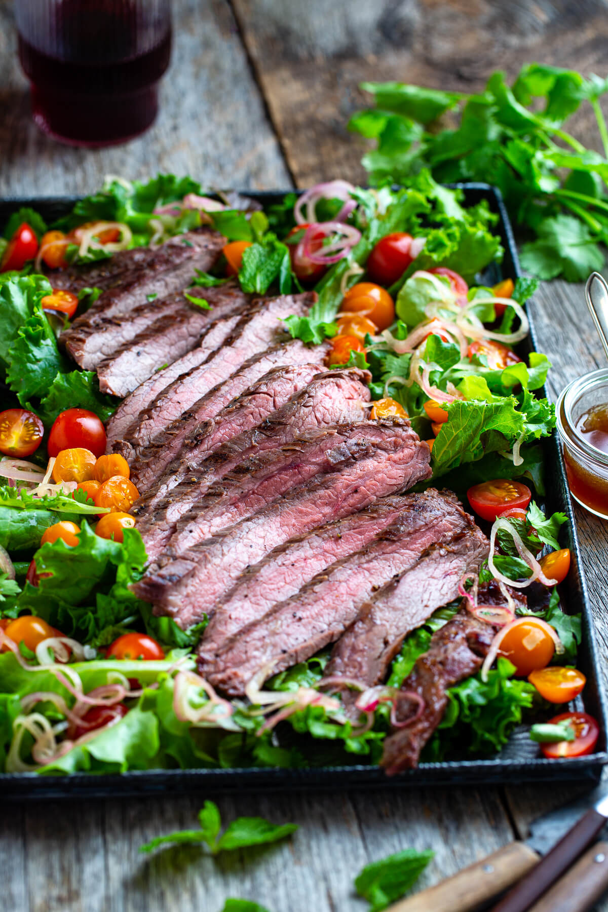 Sliced Thai beef with salad greens, tomatoes, and, fresh herbs on a metal tray with extra dressing on the side..