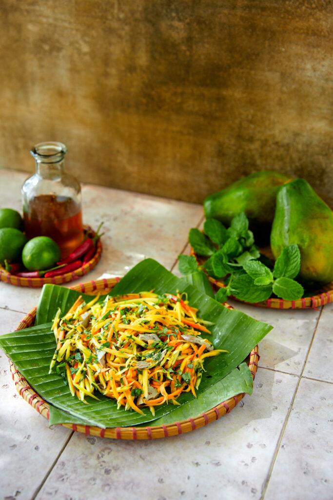 Papaya salad on some banana leaves with some mint and papaya in the background.
