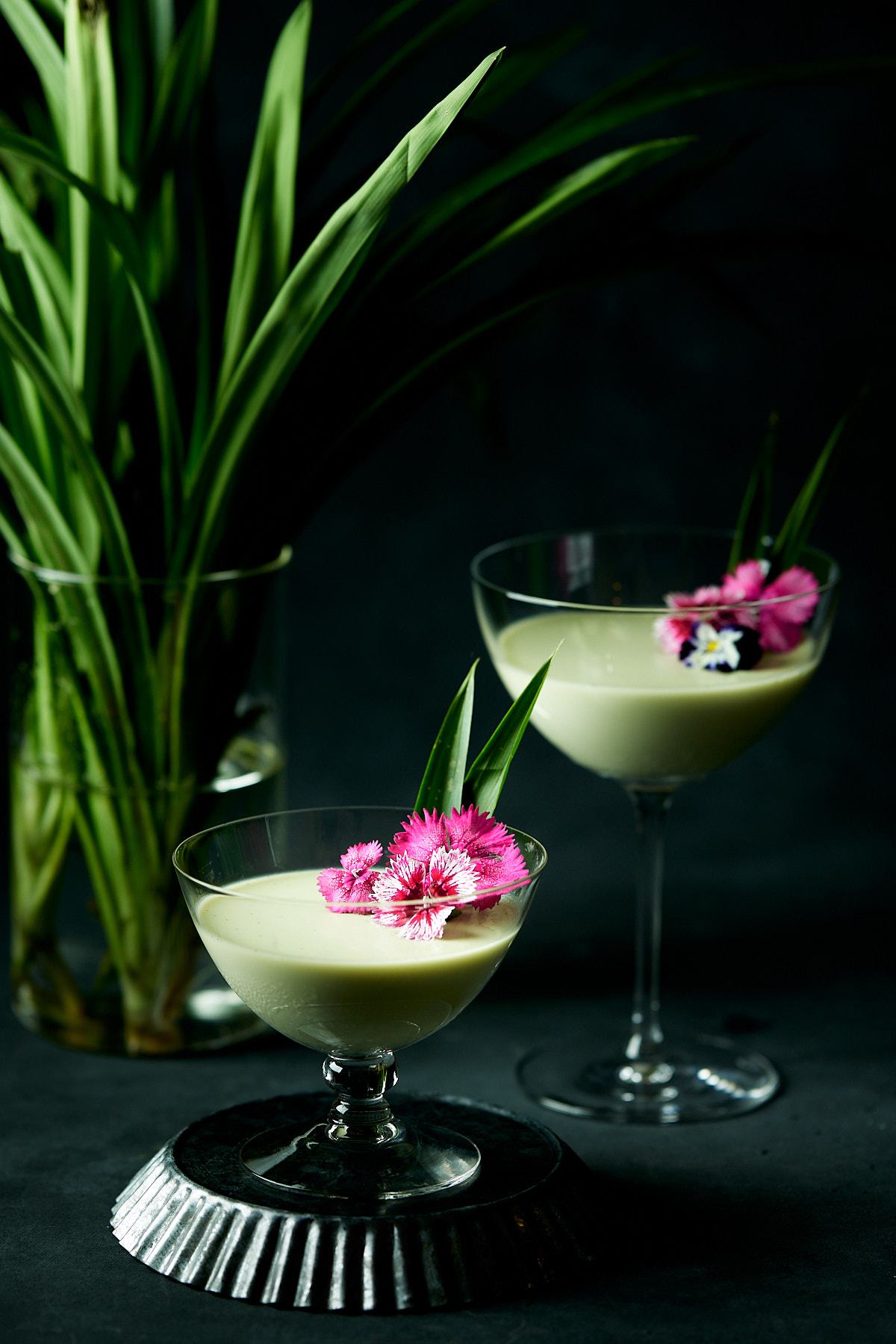 Two glasses of pandan panna cotta next to a vase of pandan leaves.