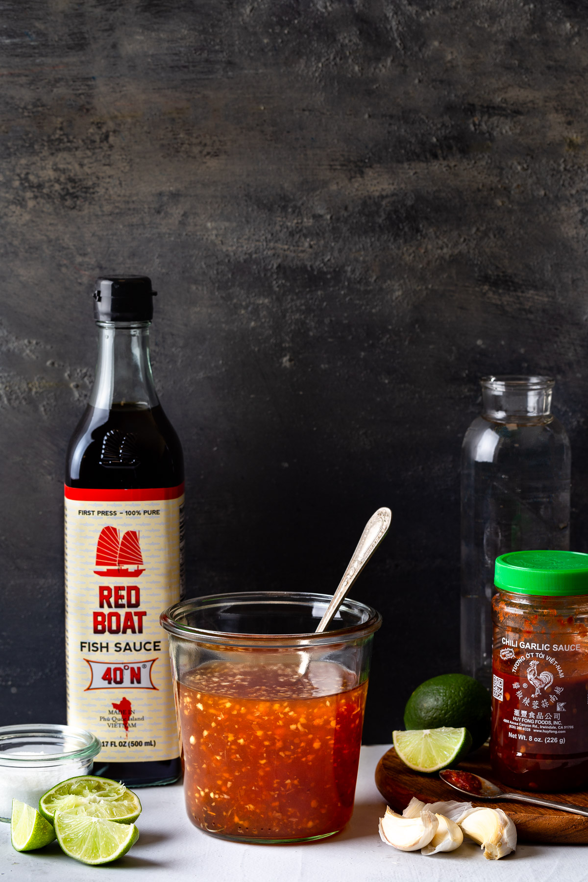 A jar of prepared Vietnamese fish sauce surrounded by a bottle of fish sauce, limes, garlic, and some chili garlic sauce.