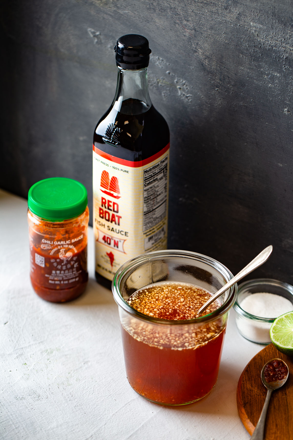 A jar of prepared Vietnamese fish sauce surrounded by a bottle of fish sauce, limes, and some chili garlic sauce.