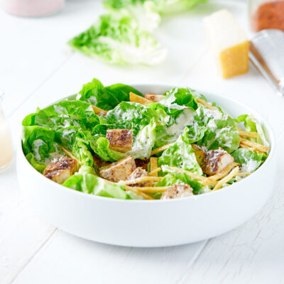 Serving bowl containing chicken caesar salad next to jar of dressing.