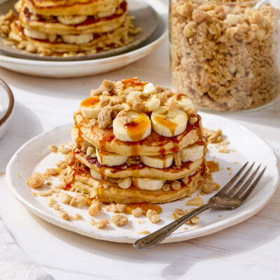 A stack of pancakes layered with banana slices and macadamia nut streusel and topped with syrup.