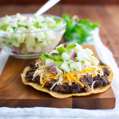 Black beans topped with cheese and green apple salsa on a tostada shell on a wooden serving board.