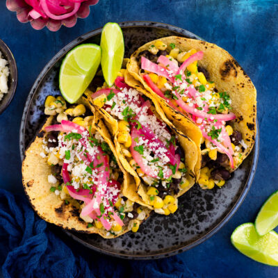 Black bean and elote tacos garnished with queso fresco, chopped cilantro, and pickled onions on a black plate.