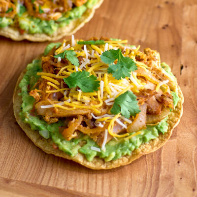 A tostada topped with guacamole, sautéed onions, spiced chicken, grated cheese, and cilantro.