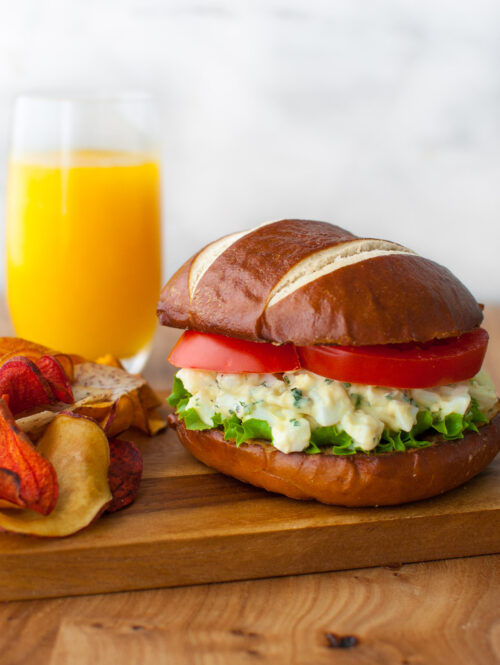 An egg salad sandwich on a pretzel roll served with chips and a glass of orange juice.