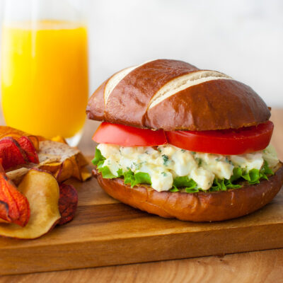 An egg salad sandwich on a pretzel roll served with chips and a glass of orange juice.