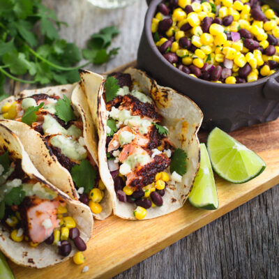 Salmon tacos next to some lime wedges and a bowl of black bean and corn relish.